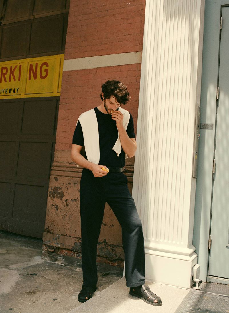A brunette male model stands in front of a brick building, wearing a black shirt and pants with a white sweater around his neck.