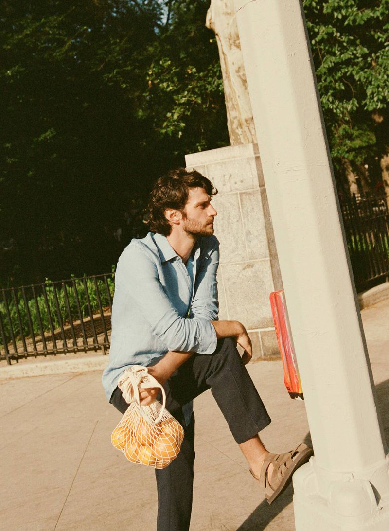 A man in a blue linen shirt and dark pants leans against a light pole outdoors holding a mesh bag full of oranges.