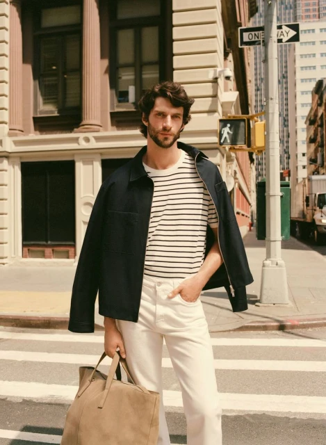 Adult man stands on city crosswalk wearing jacket, striped shirt, and white pants, holding a tan tote bag in daylight.