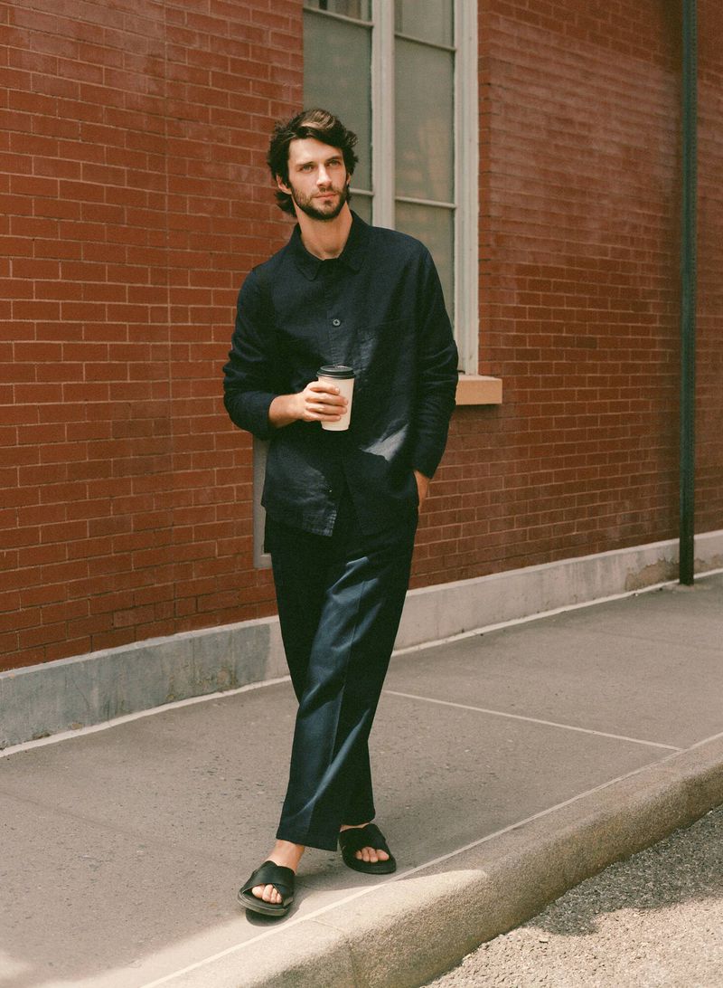 Man wearing a dark blue shirt and trousers stands against a brick wall outside while holding a disposable coffee cup in daylight.