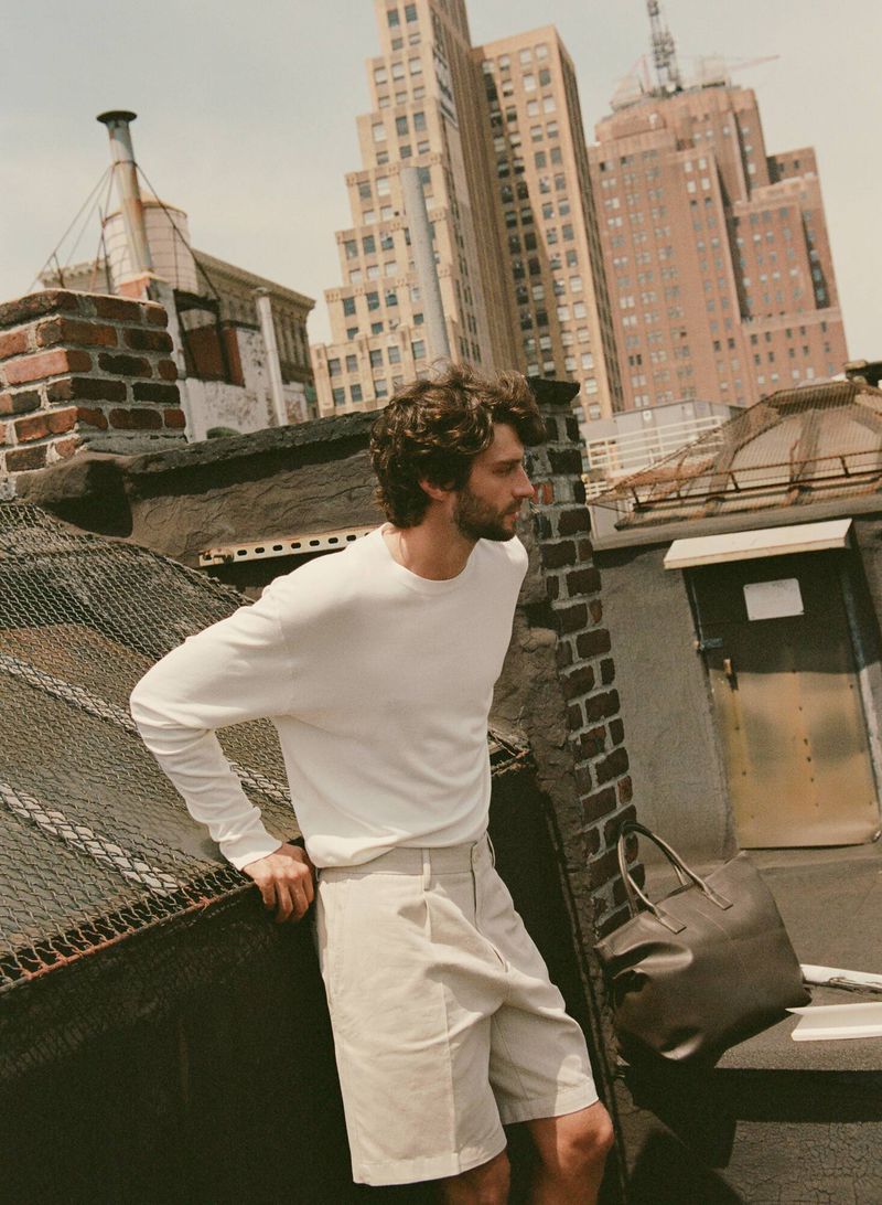 An adult male stands on a rooftop with a brick ledge, posing next to a leather tote bag, with tall buildings rising in the background.