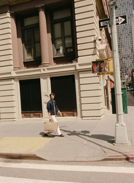 A man wearing casual clothes walks across a crosswalk carrying a tote bag in an urban setting with a building and skyscrapers in the background.