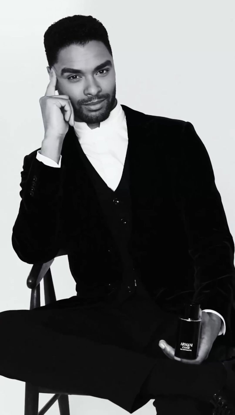 Portrait of an adult male in a black suit sitting and holding a perfume bottle against a white background in a photo studio. 