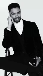 Regé-Jean Page seated in a black velvet suit holding a perfume bottle for Armani Beauty