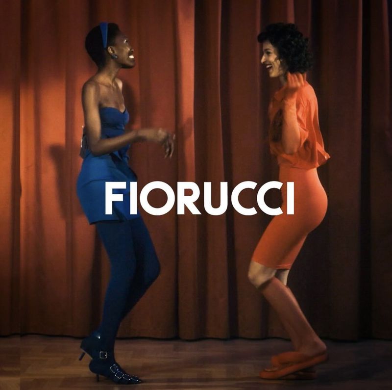 Two models pose in colorful dresses against a reddish-brown curtain backdrop, showcasing the Fiorucci brand with a playful energy.