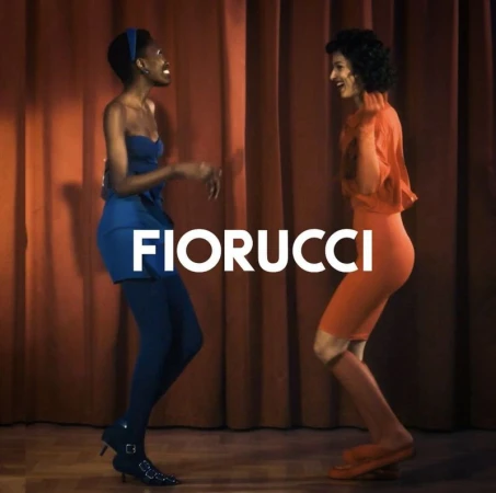 Two models pose in colorful dresses against a reddish-brown curtain backdrop, showcasing the Fiorucci brand with a playful energy.