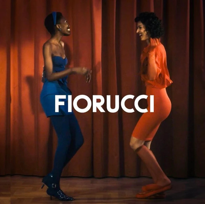 Two women laughing and dancing in front of brown curtains wearing blue and orange outfits for Fiorucci