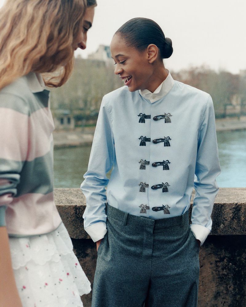 Two fashion models pose outdoors against a riverside Parisian backdrop, wearing tailored wool trousers and a light blue button-down shirt.