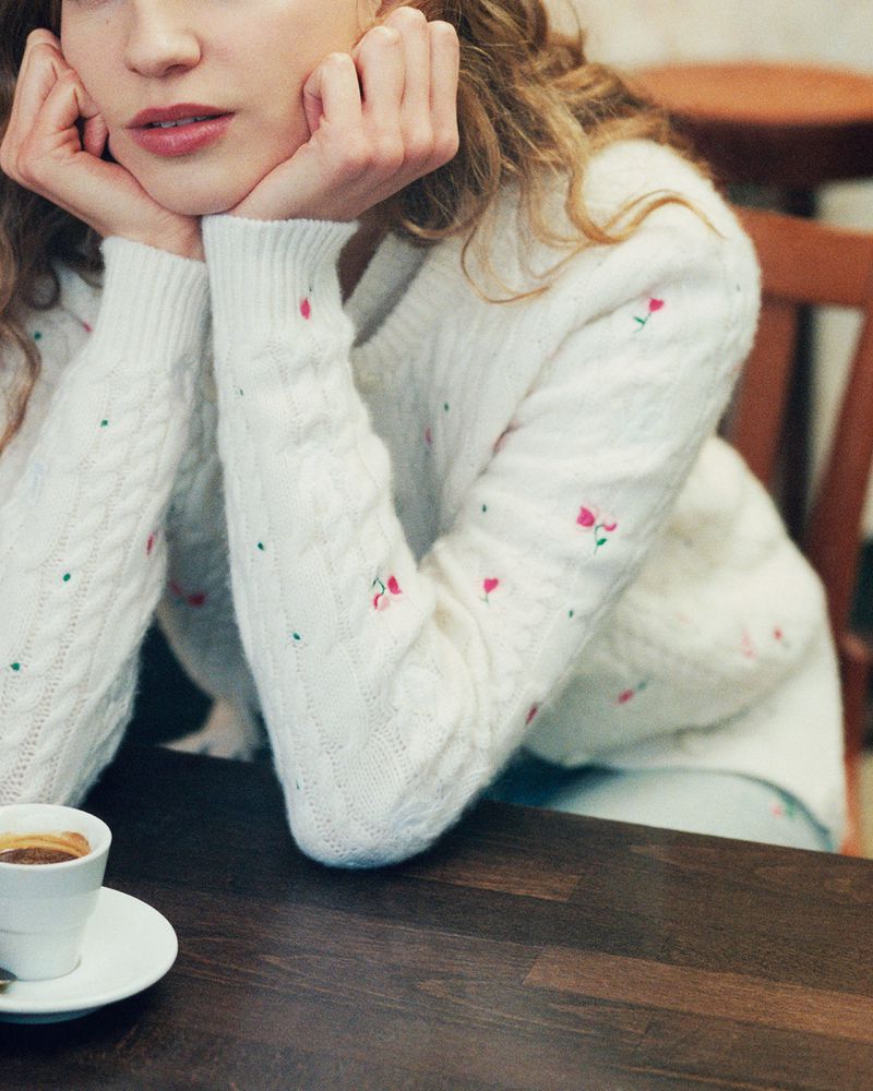 Close up of a light-skinned woman wearing a patterned knit sweater resting her chin in her hands near a cup of coffee on a table.