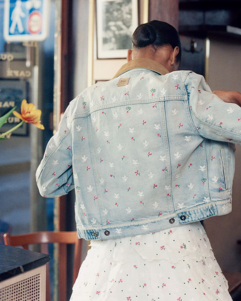 Woman wearing an embroidered denim jacket and white dress indoors. The jacket features light blue denim with floral and dove details.
