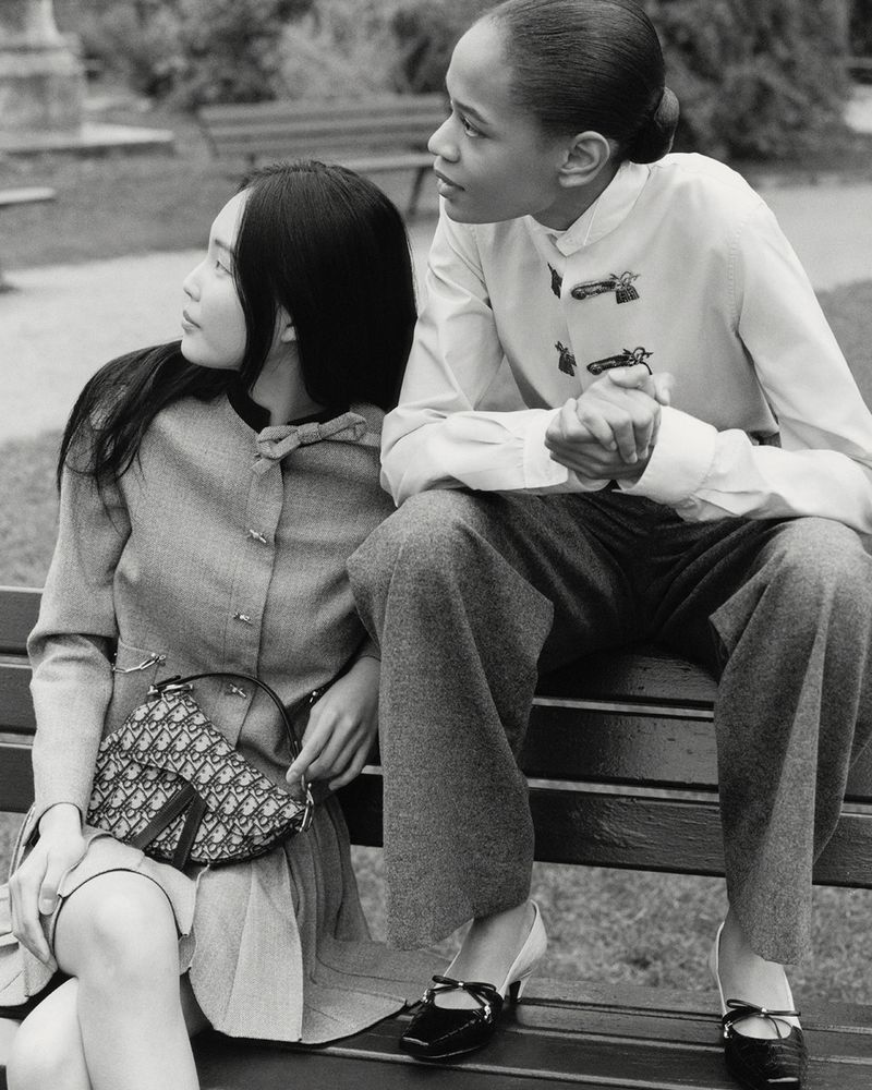 Two women in vintage fashion sit on a park bench. One carries a Dior saddlebag. Shot in black and white with a shallow depth of field.