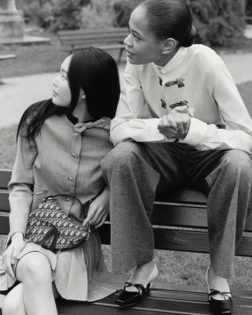 Two women in vintage fashion sit on a park bench. One carries a Dior saddlebag. Shot in black and white with a shallow depth of field.