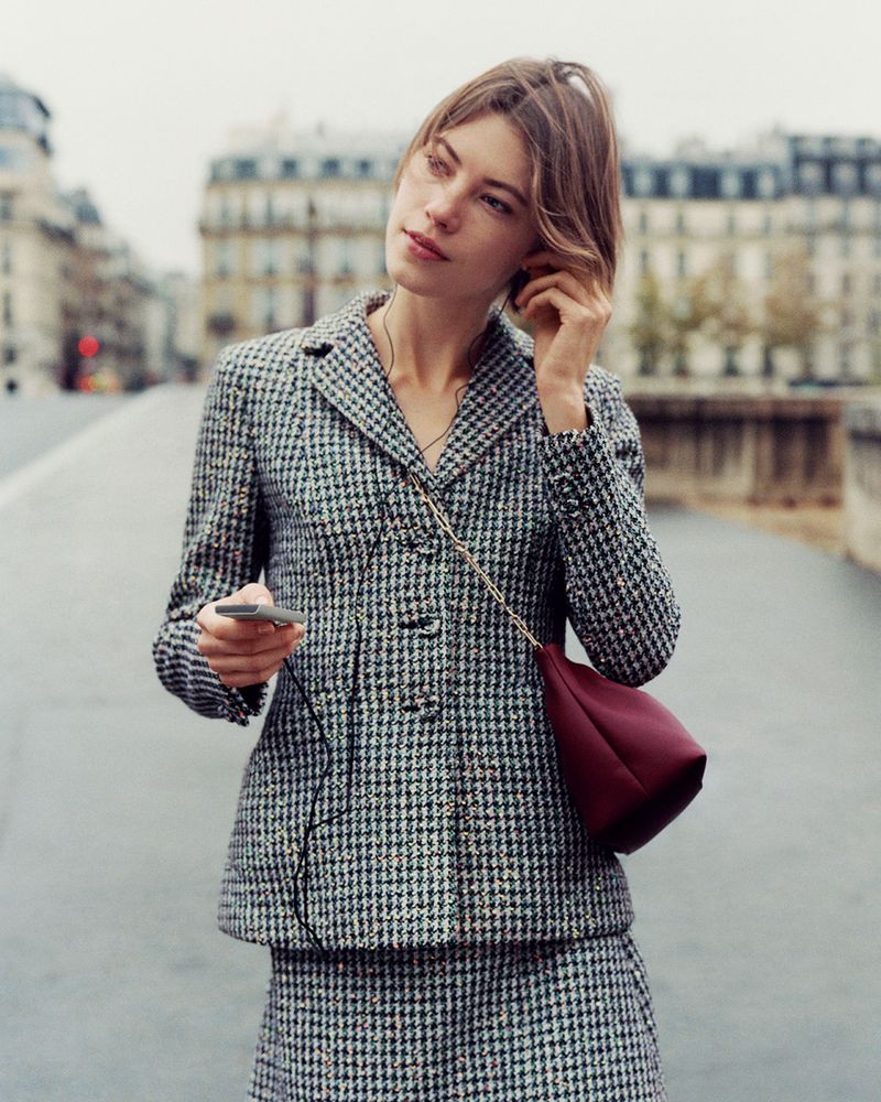 Adult woman with short hair, wearing a houndstooth jacket and skirt, stands on a bridge, holding a device with headphones in, outdoors.