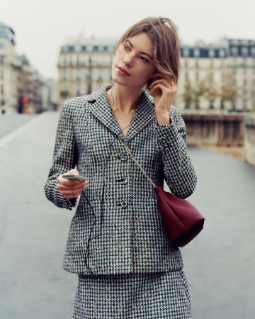 Adult woman with short hair, wearing a houndstooth jacket and skirt, stands on a bridge, holding a device with headphones in, outdoors.
