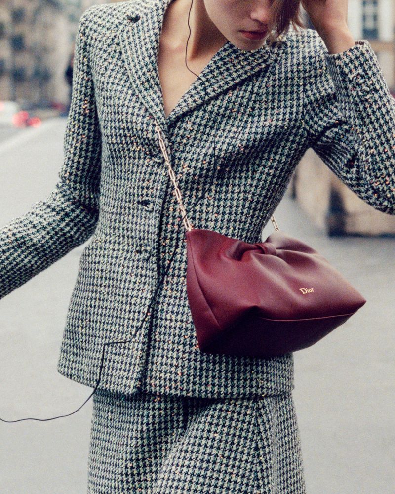 Woman in houndstooth blazer and skirt poses with a red leather Dior purse in an urban setting. Fashion, style, and luxury are visible.