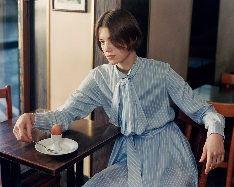 Young woman in a blue striped dress sits at a table in a cafe with a soft boiled egg in an egg cup. Interior setting, natural light.