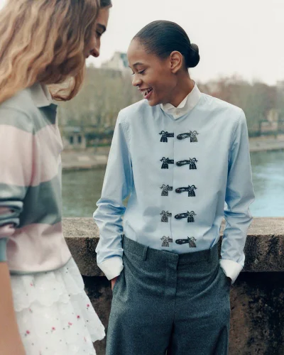 Model smiling in a light blue embroidered collared shirt and grey trousers standing next to a model on a stone river bridge.
