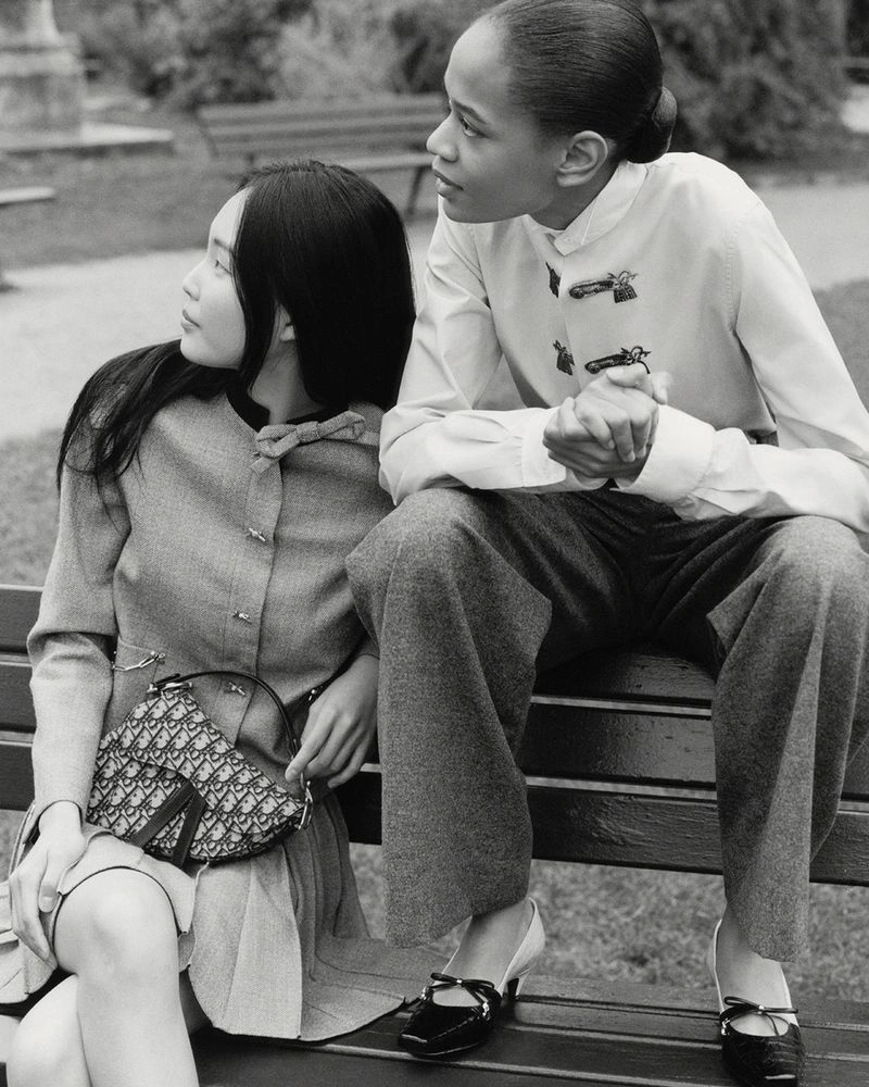 Two female models seated on a park bench wearing Dior collection attire in a black and white photograph