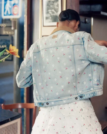 Model seen from behind wearing an embroidered light wash denim jacket with a tan collar and a white tiered skirt inside a cafe.