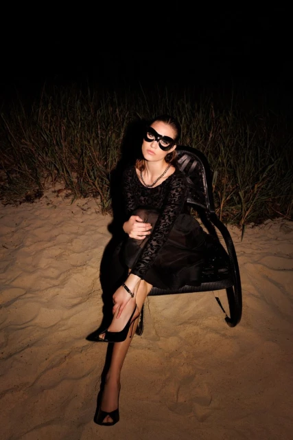 An adult female poses in a black dress and mask while sitting in a wicker chair on sand in front of green grasses at night.