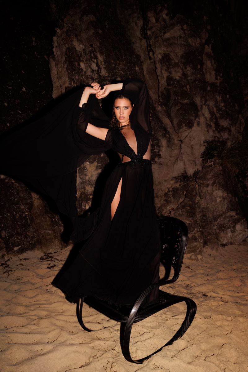 Woman in a black dress posing on a decorative chair in a rocky, cave-like setting with dramatic lighting for a fashion shot.
