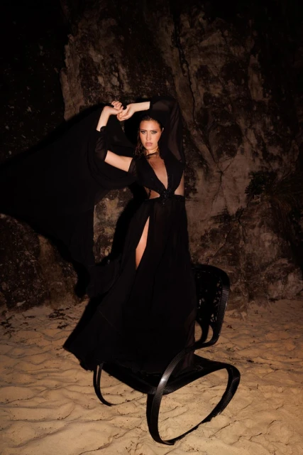 Woman in a black dress posing on a decorative chair in a rocky, cave-like setting with dramatic lighting for a fashion shot.