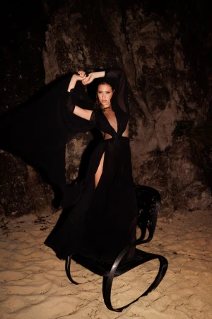 Woman in a black dress posing on a decorative chair in a rocky, cave-like setting with dramatic lighting for a fashion shot.