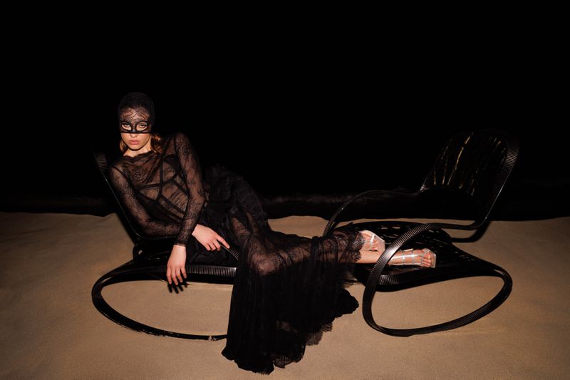 Woman in a black lace dress and mask reclines on a black chair against a dark background, creating a mysterious and fashionable scene.