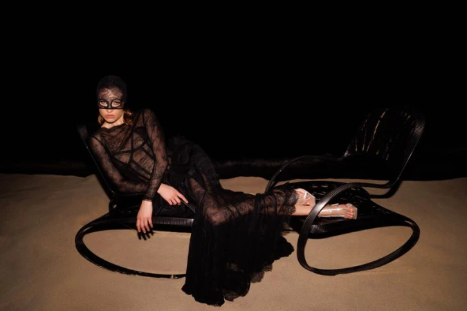 Woman in a black lace dress and mask reclines on a black chair against a dark background, creating a mysterious and fashionable scene.