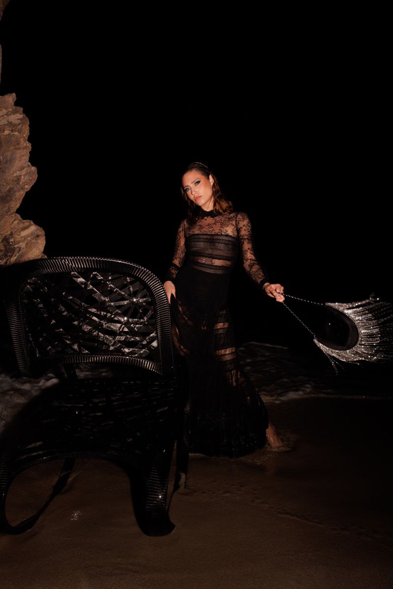 Woman in black sheer gown posing on beach at night near black wicker chair with wet sand, ocean, and cave in the dark background.