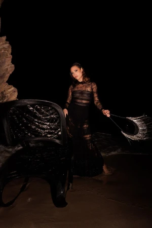 Woman in black sheer gown posing on beach at night near black wicker chair with wet sand, ocean, and cave in the dark background.
