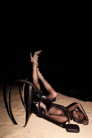 A woman in sheer black clothing reclines on a unique black chair on a sandy surface, with a dark background, wearing strappy heels.