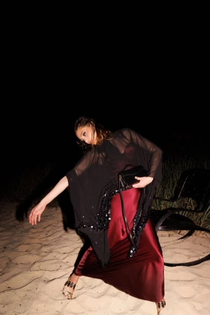 Fashionable adult female model poses on a sandy beach at night, wearing a burgundy dress, sheer shawl, and decorative sandals.