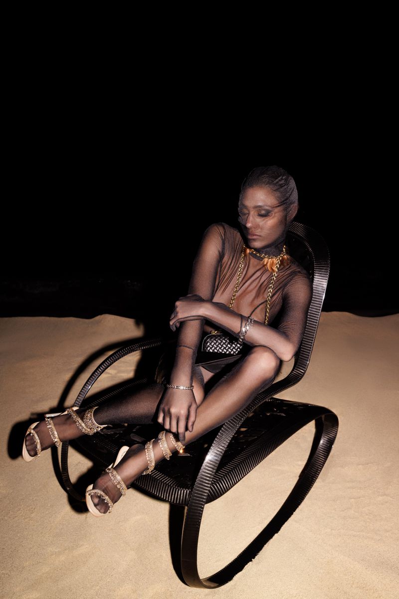 A woman sits in a modern chair on tan surface in a sheer outfit with jewelry and high heels against a dark backdrop in a studio shot.