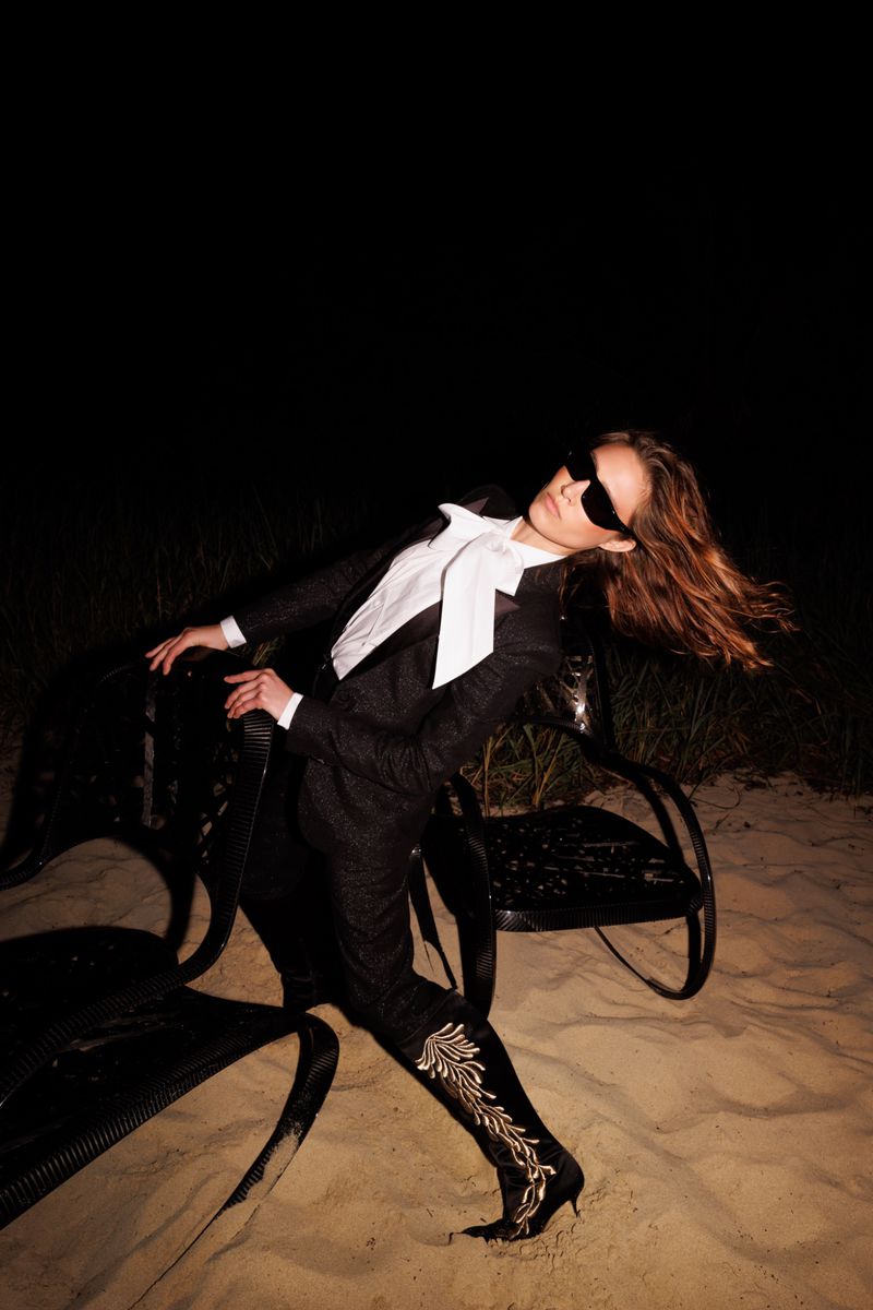 Fashion portrait of a woman in a suit leaning on a metal chair on a sandy surface with a black background.