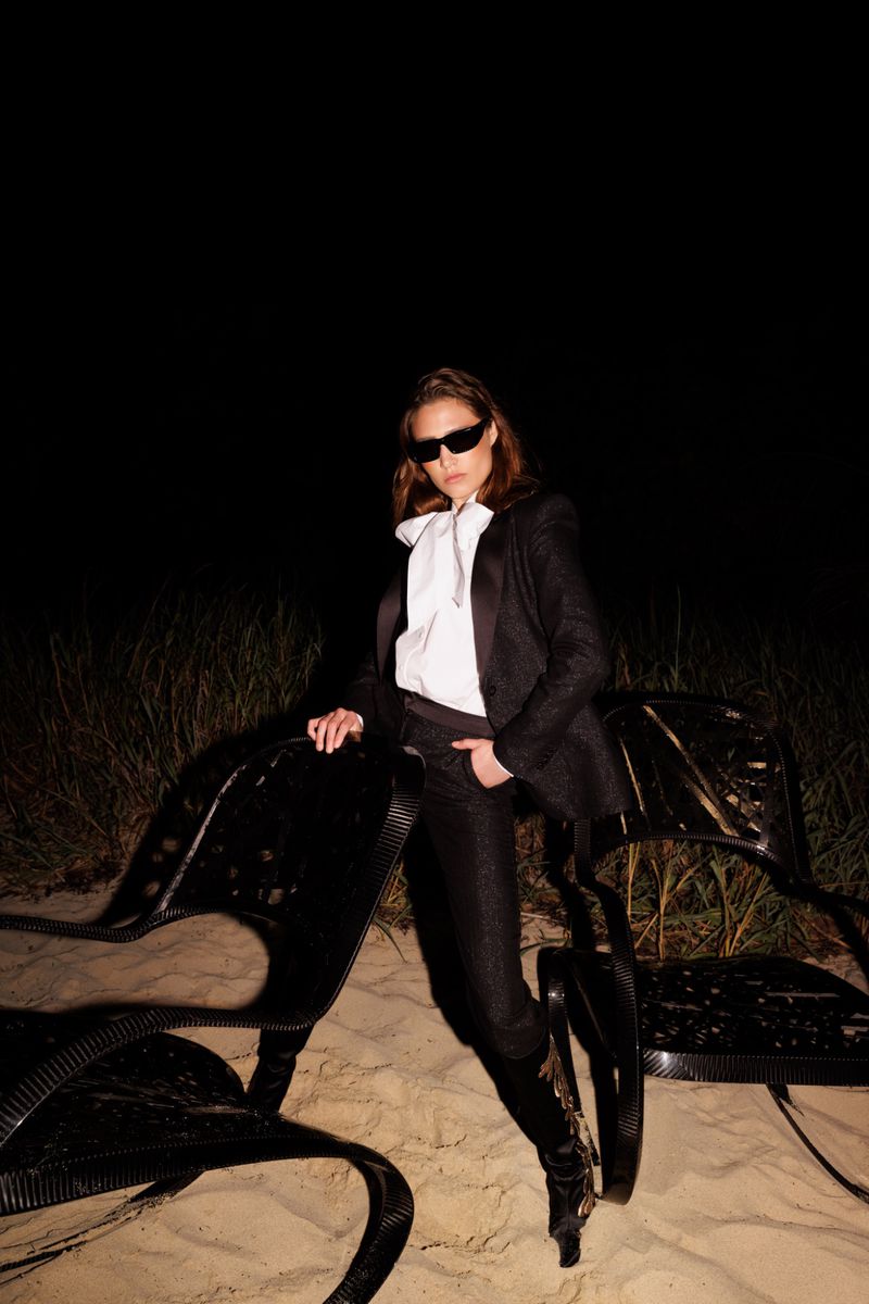 Adult female poses on a beach at night wearing a black suit, white blouse, sunglasses, and black boots, leaning on a modern black chair.