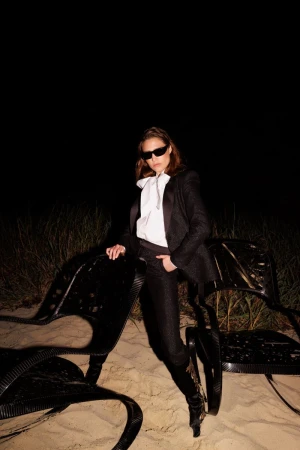 Adult female poses on a beach at night wearing a black suit, white blouse, sunglasses, and black boots, leaning on a modern black chair.