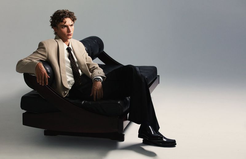 Elegant adult man with curly hair poses on a modern black leather lounge chair in a photo studio wearing a suit and tie.