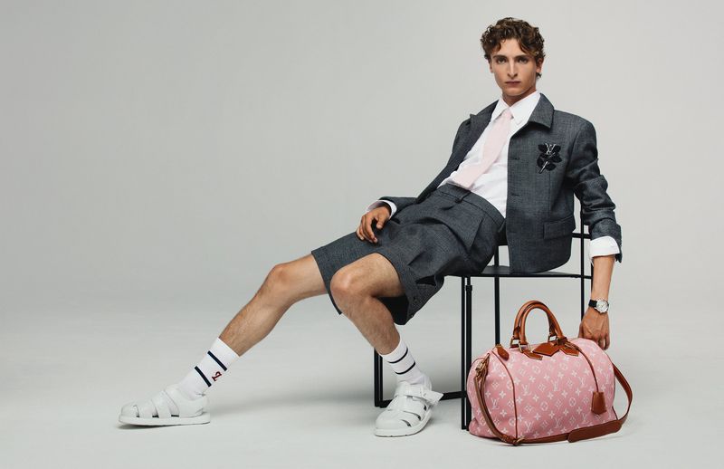 Actor Vassili Schneider seated in a grey short suit and white sandals next to a pink monogram duffel bag against a grey backdrop.
