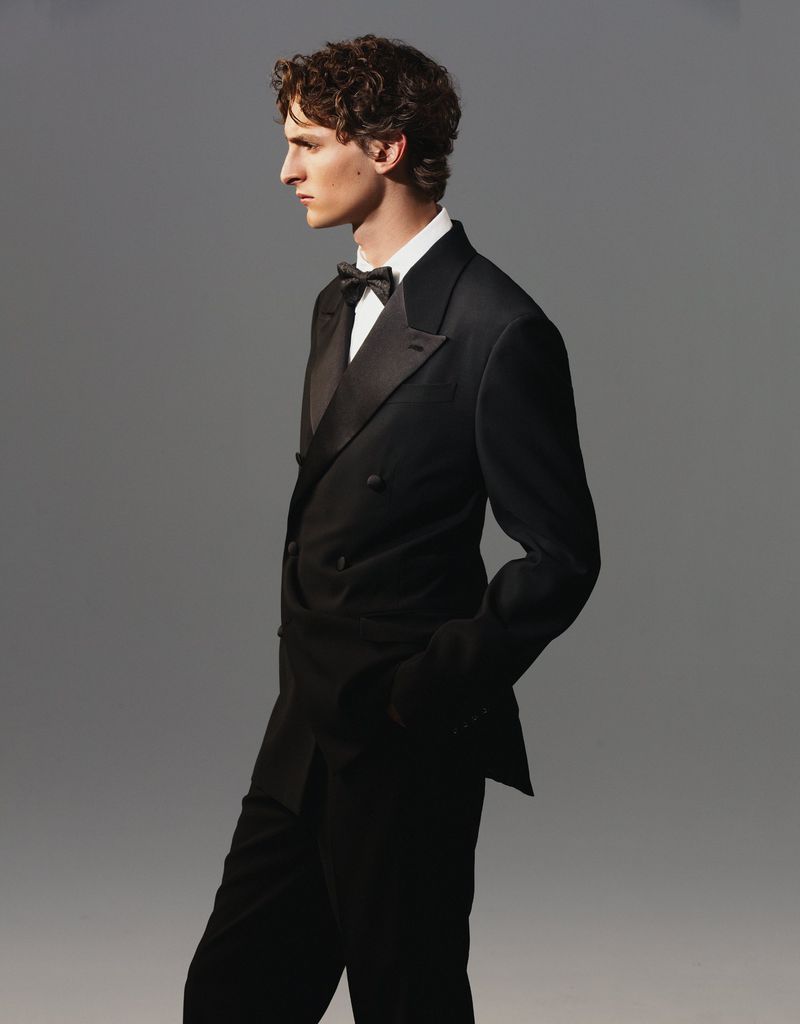 Elegant man in a classic black tuxedo with a bow tie, shot in profile against a neutral grey backdrop. Formal attire for a stylish event.