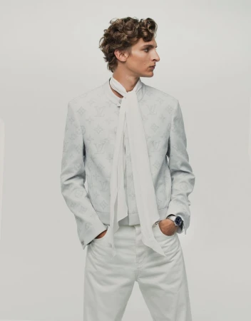 Fashionable blond male model poses in white jacket and pants with hands in pockets in a bright studio setting, looking off camera.