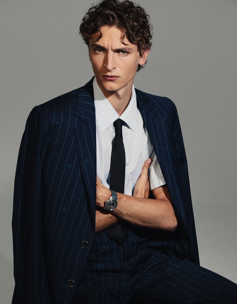 Stylish brunette man in a pinstripe suit and tie poses in a studio with crossed arms, looking confidently at the camera.