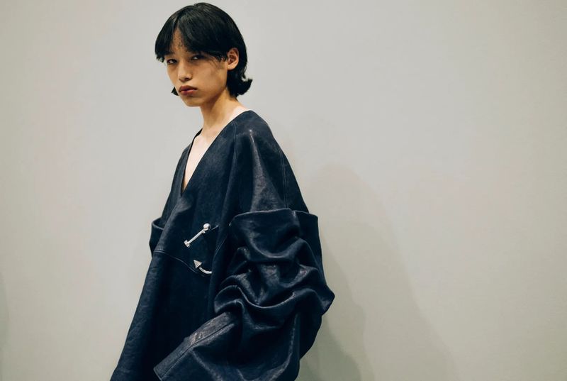 A person with dark hair poses in a dark, oversized garment with architectural shoulder detail against a neutral studio backdrop.