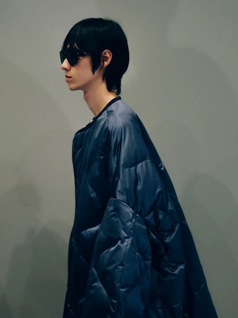 A young adult man with black hair and sunglasses stands indoors wearing a quilted blue jacket, looking to the side in a fashion shot.