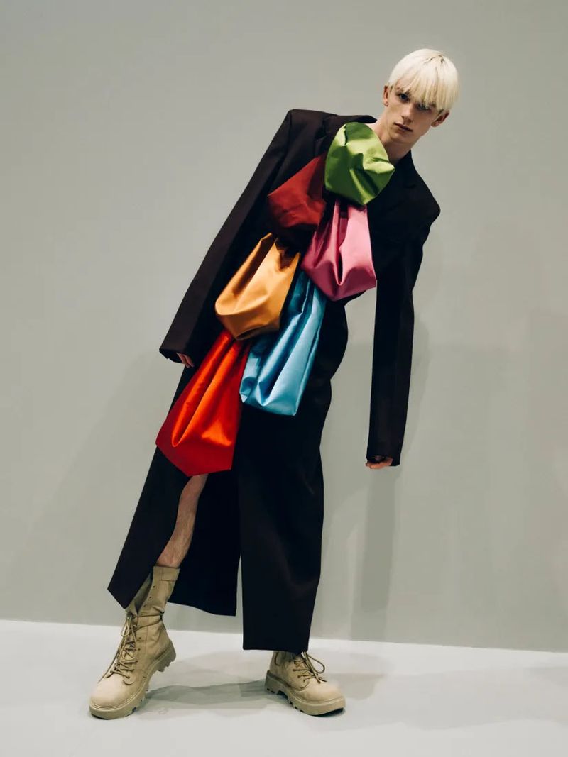 Fashion model poses in studio wearing a dark coat and colorful satin bags, accessorized with tan lace-up boots against a gray backdrop.