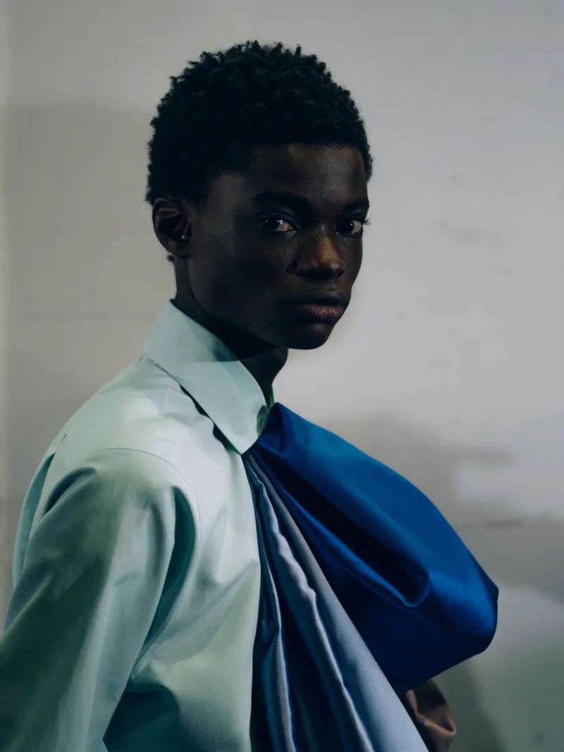 Portrait of a young Black man wearing a blue and light-green button-down shirt, posing against a neutral wall in a medium shot.