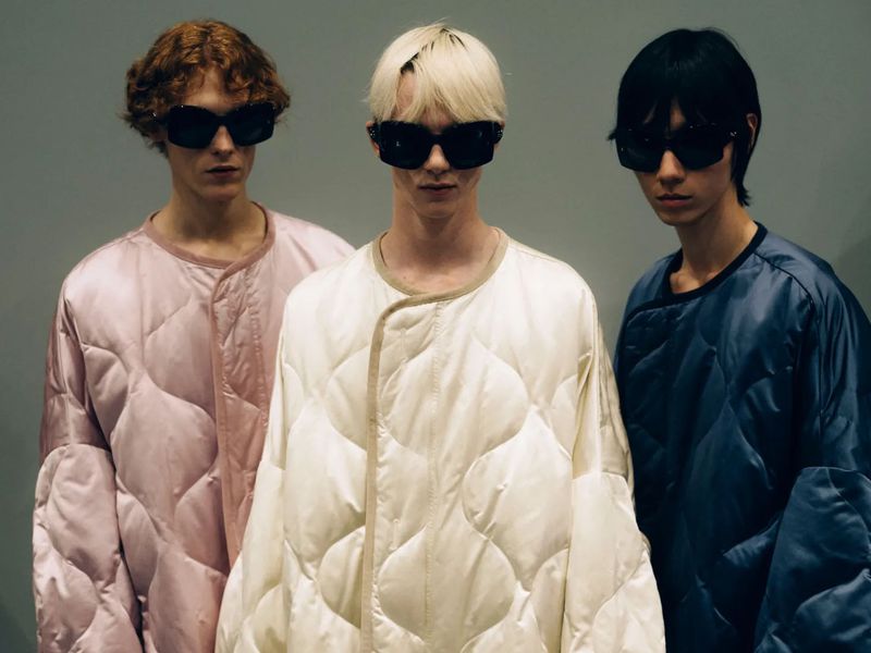 Three adult male fashion models wearing sunglasses and quilted jackets in pink, white, and blue stand indoors against a neutral background.