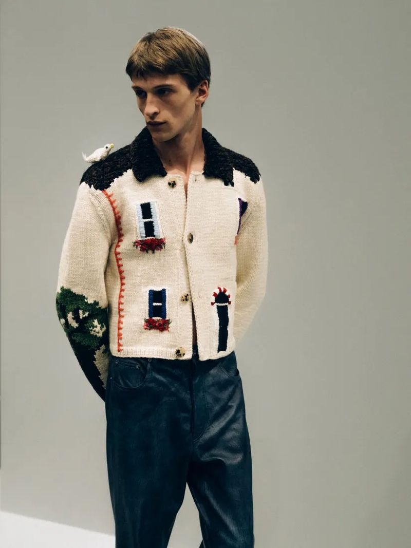 Male model in a patterned knit jacket and dark pants with a bird perched on his shoulder, posing in a bright indoor studio.
