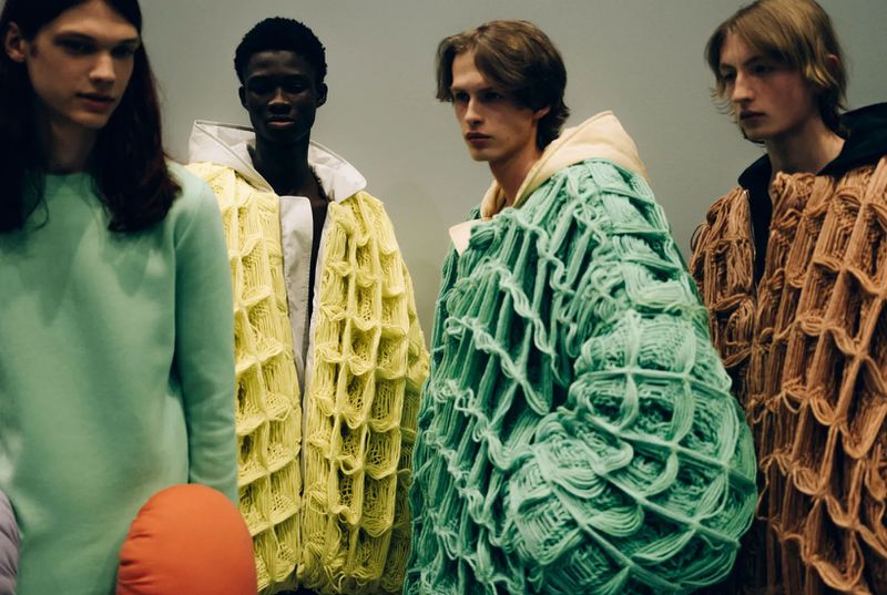 Four people model knitwear in a fashion shoot. The models are standing. One wears a light green top. Three are wearing yellow, green and brown textured jackets.