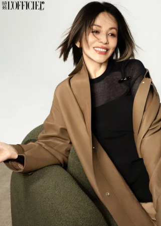 Woman smiling in a tan leather coat and black top while seated on an olive armchair against a white studio backdrop.